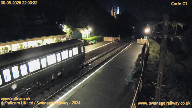 A darkened railway station platform at night, illuminated by a few bright lights. There is an empty train with illuminated windows, parked on the left side. The platform has a small shelter and a bench near the center, with some flowers and a fence at the far end. Overhead, power lines and a tall utility pole are visible. In the background, there is a faint silhouette of a landform or building on a hill. The image includes timestamps and website information at the top and bottom edges.