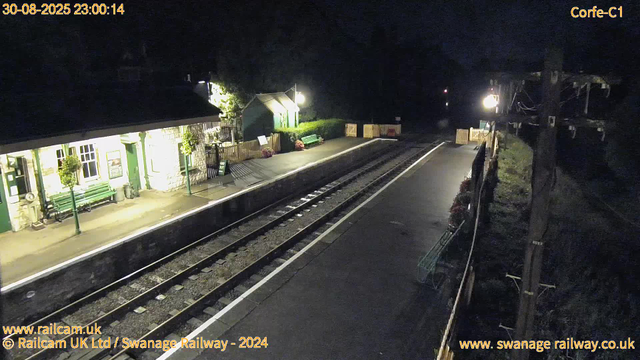 A quiet railway station platform at night, illuminated by a bright lamp. There are green-painted benches and potted plants along the station building, which has white stone walls and green doors and window frames. A small green shelter is visible in the background. The railway tracks run down the middle, empty at this moment. On the right side, there is a tall wooden telegraph pole with electrical wires and a small flower bed at its base. No people are present on the platform. The date and time are displayed at the top and bottom of the image, and there is text indicating the railway and website information.