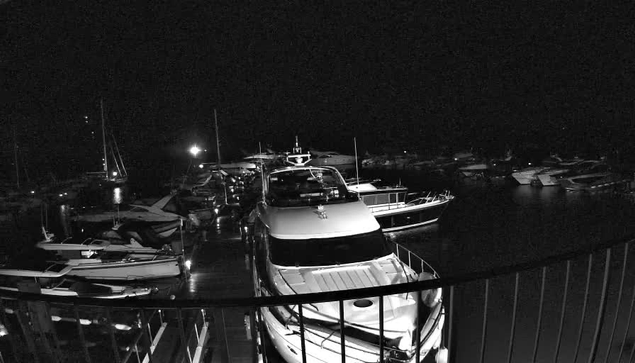 A nighttime scene at a marina with several boats docked in the water. The foreground shows a large yacht facing the camera, with a railing in front. In the background, smaller boats and sailboats are moored, with dark water and a dark sky surrounding the marina. Some lights are faintly visible, reflecting on the water.