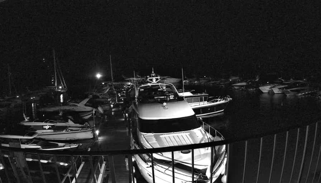 A black and white image of a harbor at night showing multiple boats docked in the water. In the foreground, a large yacht is seen from above, with a railing visible at the bottom edge of the image. The background includes more boats and faint lights reflecting on the water, creating a tranquil nighttime scene.