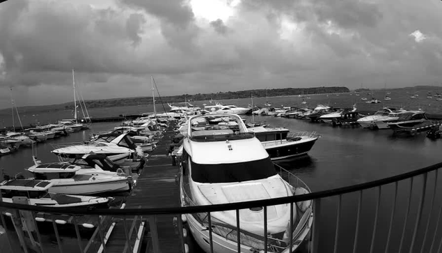 A black-and-white photo taken from a webcam shows a marina filled with various boats and yachts docked along the pier. A railing is visible in the foreground, curving across the bottom of the image. The sky above is cloudy and overcast, with a distant shoreline visible in the background.