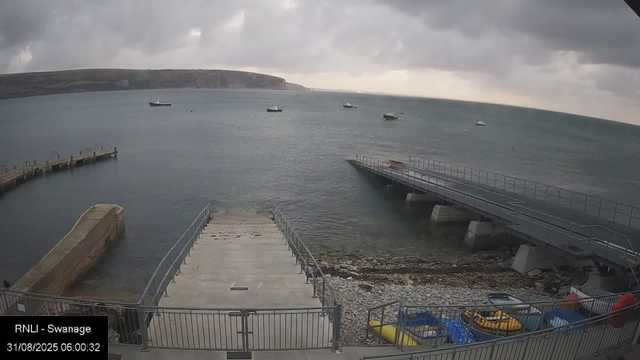 A view of a cloudy sky over a calm sea with several boats anchored in the water. In the foreground, there's a small dock with metal railings and a concrete ramp leading into the water. To the right, there's a pier extending into the sea. At the bottom, there are colorful kayaks and small boats stored on a boat rack. The image has a timestamp indicating it was taken on August 31, 2025, at 6:00 am, and is labeled "RNLI - Swanage."