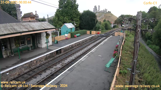 A small train station platform with a single set of railway tracks runs through the scene. To the left, there is a stone station building with a tiled roof, flower baskets hanging outside, and green benches for waiting passengers. A small, green, metal shed and colorful flower arrangements are situated near the platform. To the right, power lines and poles run parallel to the tracks, with some greenery and trees in the background. In the distance, on a hill, the ruins of a castle are visible, set against a partly cloudy sky. The timestamp indicates early morning, August 31, 2023.