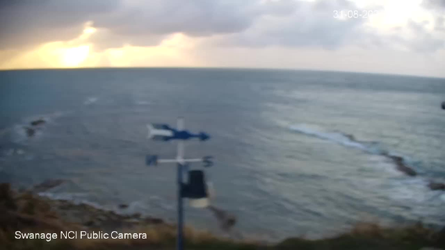 A view of the ocean from a webcam, showing a cloudy sky with the sun partially obscured by dark clouds. The coastline is visible at the bottom of the image with some rocks and waves crashing against it. An anemometer (weather instrument) is in the foreground, and the timestamp "31-08-2023" is displayed in the upper right corner.