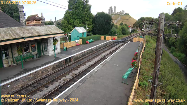 A small train station platform with a stone and wooden building on the left, decorated with hanging flower baskets. There are green benches along the edge of the platform, some with flower pots nearby. A green wooden shed is behind the benches, surrounded by a yellow picket fence. The platform runs parallel to two railway tracks, which extend into the distance. On the right side, there are wooden utility poles with wires, and a pathway with a green fence and red flower pots. In the background, there is a hill with an ancient castle ruin on top, surrounded by greenery. The time and date displayed are 07:00:26 on August 31, 2025.