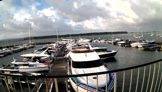 A marina with multiple boats docked along a wooden pier extending into the water. The boats vary in size and design, and some are covered while others are open. The sky overhead is partly cloudy, with patches of sunlight breaking through the clouds, casting reflections on the water. In the background, there is a distant shoreline with trees.