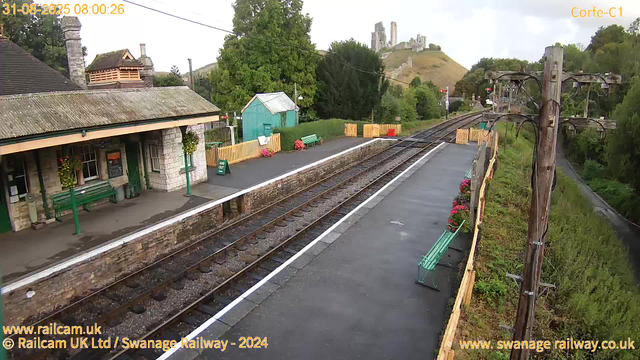 A small train station platform with a stone building on the left, decorated with hanging flower baskets and green benches. There are yellow fences and flower pots near the platform edge. A set of train tracks run through the center, with a wooden utility pole on the right side supporting overhead electrical wires. In the background, a hill with the ruins of a castle or large structure is visible atop a hill, surrounded by trees and greenery. The sky is partly cloudy, and the scene appears calm.