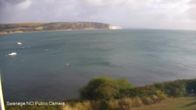 A view of a large body of water with several small boats floating on the surface. In the background, there is a coastline with a white chalk cliff and some green hills. The sky is mostly cloudy with some sunlight breaking through. The image appears to be captured from a webcam overlooking the coastal area. The timestamp on the top right reads "31-08-2023 06:00:41," and the bottom left indicates "Swanage NCI Public Camera."