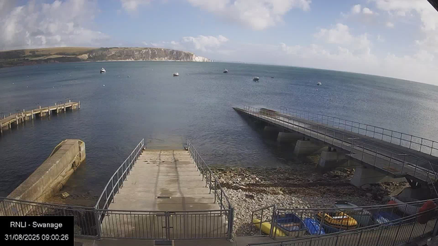 A view of a calm seaside scene with a concrete pier extending into the water. To the left, a small, angled platform is visible, with metal railings along the edges. Several boats are anchored in the distance across the water. In the background, white cliffs rise along the horizon under a partly cloudy sky, with patches of sunlight illuminating the scene. There are some small vehicles and equipment parked near the shore at the bottom of the image.