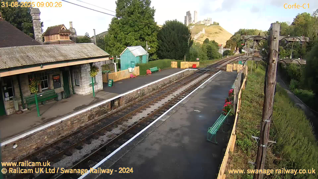 A small rural train station platform with a stone building on the left, decorated with hanging flower baskets. The platform has green benches and a "Way Out" sign. There are multiple railroad tracks running parallel, with a wooden fence and potted flowers along the edge. In the background, there are trees, a green shed, some colorful containers, and a hillside castle or ruin atop a hill. An overhead electric line and pole are visible on the right side. The sky is partly cloudy with periods of sunlight.