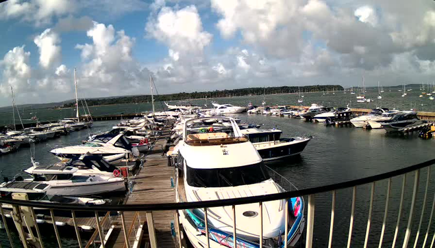 A marina filled with numerous boats and yachts docked at wooden piers, with a cloudy sky overhead and a distant landmass visible across the water. A metal railing is in the foreground, partially framing the scene.