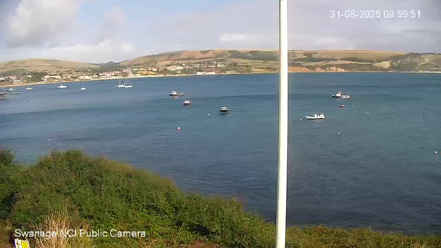 A view of a harbor with several small boats floating on calm water. In the background, there are green hills and a town with buildings along the shoreline. The sky is partly cloudy, and the image has a timestamp in the upper right corner. The bottom left corner has the text "Swanage MC Public Camera."