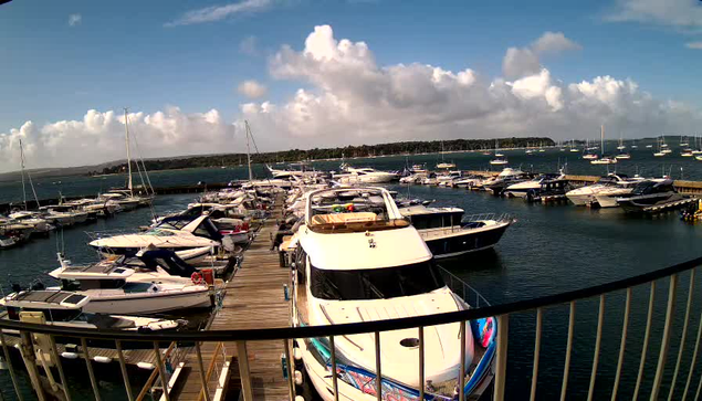 A marina filled with various white and dark-colored boats and yachts docked along wooden piers. The sky is partly cloudy with a mix of white clouds and blue sky, and the water is calm. A black railing is visible in the foreground.