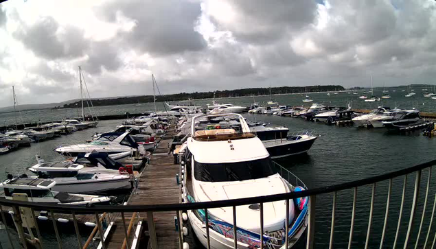 A marina with numerous boats docked along a wooden pier, extending from the foreground into the distance. The boats vary in size and style, mostly motorboats and yachts, some with canopies. The water is calm, and the sky is partly cloudy with patches of sunlight breaking through. In the background, there is a shoreline with trees and land extending across the horizon. A black railing curves along the foreground, partially framing the scene.