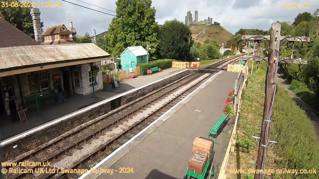 A small train station platform with a stone building on the left, featuring a tiled roof and a chimney. There are a few people near the entrance. The platform has a green bench, a small turquoise shed, and flower arrangements in planters along the edge. Railroad tracks run parallel to the platform, extending into the distance. To the right of the tracks, there are wooden utility poles with wires, and some trees and bushes. On the hill in the background, the ruins of a castle are visible atop the landscape. The sky is partly cloudy with patches of sunlight. The timestamp indicates the date is August 31, 2025, at 11:00 AM.