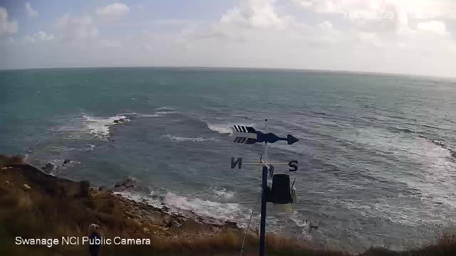 The image shows a view of the ocean from a coastal area with a rocky shoreline in the foreground. There is a weather vane with an arrow and directional markers (N, S, E, W) mounted on a pole near the bottom of the image. The sky is partly cloudy, and the water appears to be slightly choppy with small waves. The horizon line stretches across the image, separating the sea from the cloudy sky. The text "Swanage NCI Public Camera" is visible in the bottom left corner.