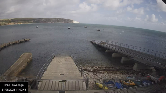A view of a harbor with two piers extending into the water. Several boats are anchored in the sea, and a distant shoreline with cliffs and hills is visible under a partly cloudy sky. In the foreground, there are colorful kayaks or small boats stored behind a fence, and the scene appears to be taken from a webcam at Swannage, as indicated by text at the bottom left corner.