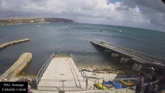 A coastal scene with a small harbor, featuring a concrete slipway leading into the water, surrounded by metal railings. Several boats are anchored in the water, and a large boat is visible near the horizon. The sky is partly cloudy with patches of sunlight illuminating the landscape. There are cliffs and rolling hills in the background, and a few people are walking along the promenade near the railings in the foreground.
