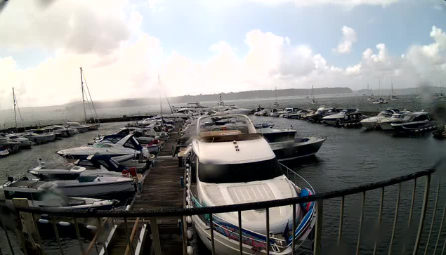 A marina filled with numerous white and dark-colored yachts and boats docked along wooden piers. Several sailboats are anchored further out in the water, and the sky is partly cloudy with patches of blue. The water appears calm, and the view extends to a distant shoreline with a few small structures or trees. A black metal railing is visible in the foreground.