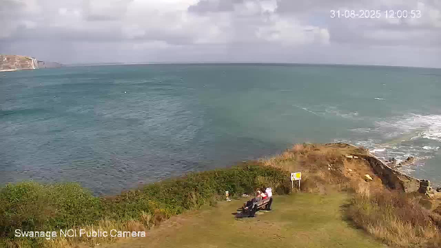 A view of the ocean with waves gently crashing onto a rocky shoreline. In the foreground, there is a grassy area with a wooden bench where three people are sitting, facing the sea. The sky is partly cloudy, and there is a coastline visible in the distance on the left side of the image. The timestamp on the top right corner reads "31-08-2025 12:00:53" and the bottom left corner displays "Swanage NOI Public Camera."
