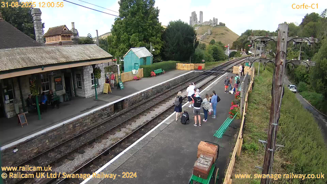 A small train station platform with several people waiting and some walking, surrounded by greenery and colorful flowers. In the background, there is a traditional stone building with a covered waiting area, benches, and signs. A group of passengers with luggage are gathered near one end of the platform, and some are engaged in conversation. There are railway tracks running through the scene, with a hill featuring an old castle ruin visible in the distance. Additionally, a wooden electricity pole with wires is on the right side of the image, and a car is driving on a road beside the station. The timestamp on the image indicates the date and time as August 31, 2025, at 12:00:56.