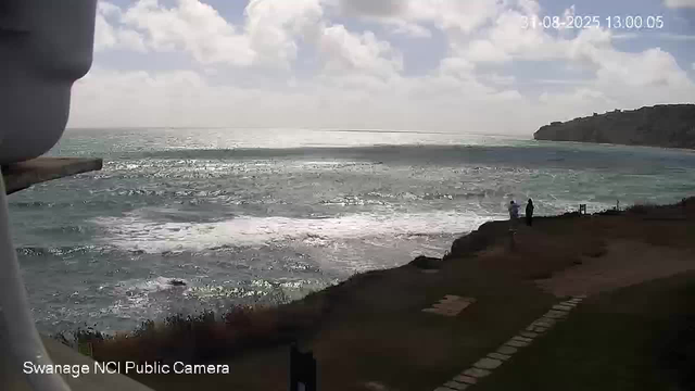 A view of the ocean with waves crashing onto a rocky shoreline. The sky is partly cloudy with sunlight reflecting off the water. Two people are standing near the edge of the land, looking out at the sea. There is a grassy path with stepping stones leading towards the shoreline on the right side of the image. A hill covered with greenery is visible in the background on the right. In the top right corner, there is a timestamp. The bottom left corner displays the label "Swanage NCI Public Camera."