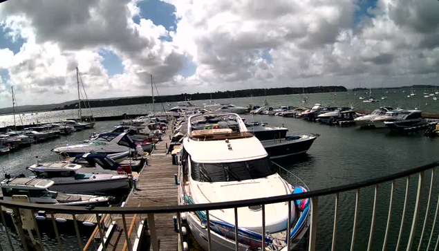A marina filled with numerous sailboats and motor yachts docked along wooden piers, with some boats covered with tarps. The sky is partly cloudy with patches of blue, and the water appears calm. A black metal railing is in the foreground, curving across the bottom of the image. In the background, there is a distant shoreline with trees and a cloudy sky overhead.