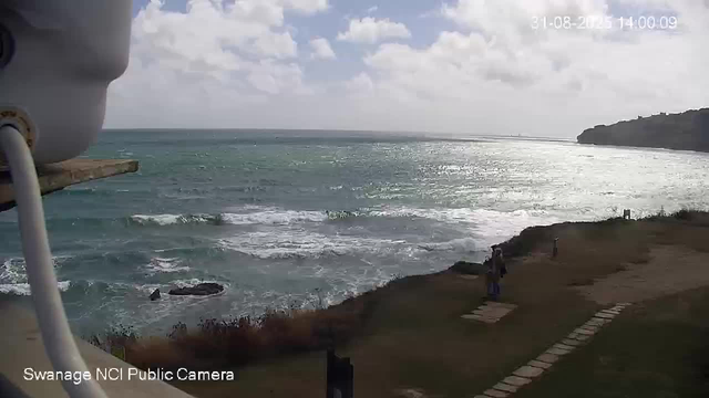 A view of a coastline with the ocean extending into the distance, waves crashing against the shore, and a partly cloudy sky overhead. There are a few people walking along a grassy path near the water, and a coast with a small hill or headland on the right side. The photo was taken during daytime.