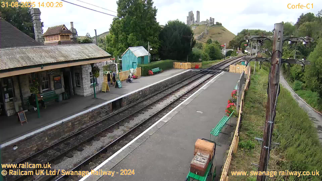 A small train station platform with a building on the left side, featuring a sloped roof and stone walls. There are a few people and a child in a stroller waiting on the platform. A blue shed, green benches, and potted plants with flowers are also visible. Railway tracks run through the center, curving into the distance with a hilltop castle ruins in the background. On the right side, there are overhead electrical wires supported by wooden poles, and greenery along the scene. The sky is mostly cloudy. The image includes timestamp and website information in the top and bottom corners.