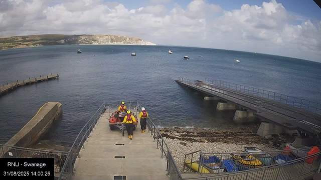 A view of a seaside harbor with a concrete dock extending into the water. Four rescue personnel, dressed in yellow and red gear, are walking on the dock toward a small boat. To the right, there is a ramp leading into the water, supported by concrete pillars. Several small boats are anchored in the water, and a cliff with grassy fields is visible in the distance under a partly cloudy sky.