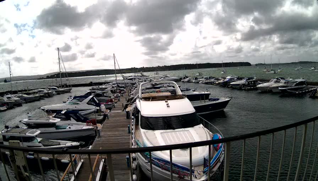 A marina filled with various boats and yachts docked along wooden piers. The water is calm, and several sailboats with masts are visible in the background. The sky is partly cloudy with patches of bright light breaking through the clouds. In the foreground, a large white yacht is prominently parked at the dock, with the bow facing towards the camera. The scene is peaceful and scenic, with a distant landmass visible on the horizon.