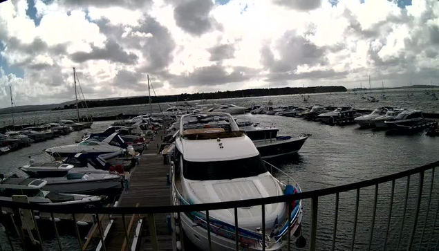 A marina filled with numerous boats and yachts docked along a wooden pier, with some boats facing towards the camera and others extending into the water. The sky is cloudy with patches of sunlight breaking through, and there is a distant shoreline or landmass visible on the horizon.