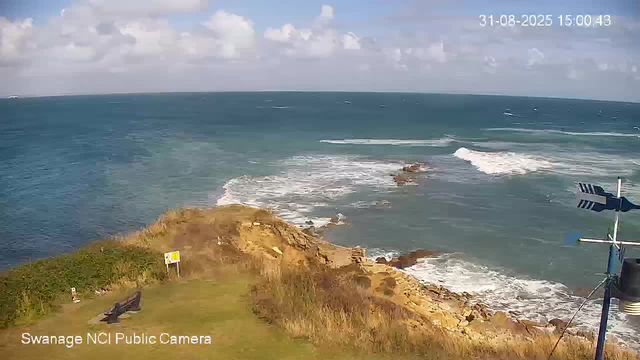 A scenic view of a coastal landscape with a rocky cliff extending into the ocean. The sea has gentle waves, and the sky is partly cloudy with some scattered clouds. In the foreground, there is a grassy area with a bench and a small sign, along with a weather vane or anemometer on the right side. The timestamp on the top right indicates the date and time as August 31, 2025, at 3:00 PM. The image is from a webcam operated by Swanine NCI Public Camera.