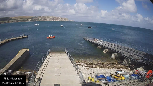 A view of a harbor with a concrete ramp leading into the water, flanked by metal railings. Several boats and yachts are floating in the water, and a small orange rescue boat with four people wearing yellow safety gear is moving across the harbor. In the background, there are cliffs and a partly cloudy sky. At the bottom left corner, there is text that reads "RNLI - Swanage" and a date and time stamp "31/08/2025 15:00:46".