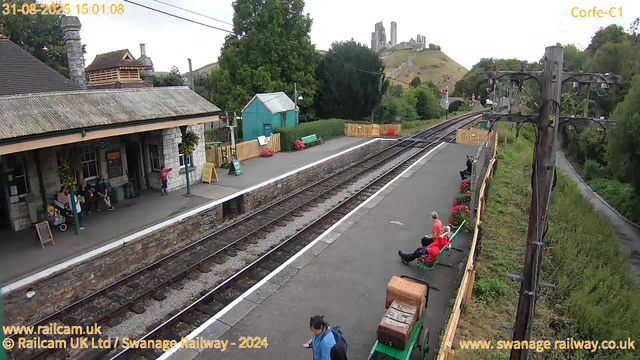 A small train station platform with a stone building on the left, where several people are sitting or standing. Some are waiting near the entrance, and one person is looking at their phone. On the right side of the platform, a few people are sitting on green benches, with pink flowers in pots along the fence. There are railway tracks running through the middle, with a railway line extending into the distance. In the background, green trees, a small turquoise shed, and a scenic hillside with a castle ruin perched on top can be seen. The sky is partly cloudy, and there are overhead electrical lines supported by wooden poles. The timestamp indicates the photo was taken on August 31, 2025, at 3:01 PM.
