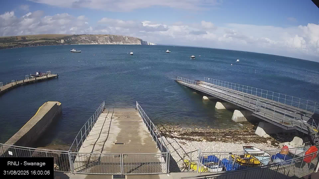 A scenic view of a harbor with a concrete and metal pier extending into the calm blue water. Several small boats are anchored in the distance, floating on the water. The sky is partly cloudy with patches of sunlight, and a hillside with cliffs and greenery is visible on the horizon.