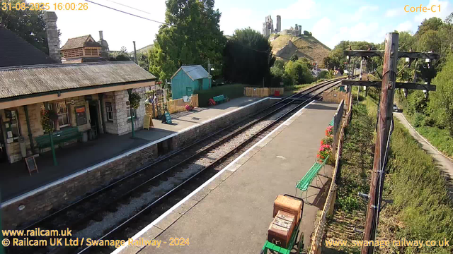 A small train station platform with a stone building featuring a sloped roof and hanging flower baskets. There are a few benches, a small green shed, and a fenced area with colorful flowers. Overhead, electric train wires are visible, supported by wooden poles. In the background, a hill with ruins or old structures and lush green trees are visible under a partly cloudy sky. The timestamp indicates it is late afternoon on August 31, 2025.