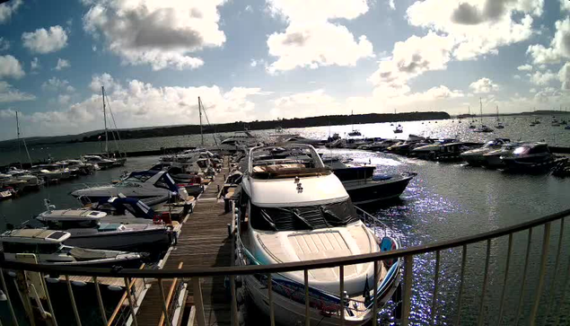 A marina filled with numerous docked boats and yachts. The water is calm and reflects the sunlight, with a partly cloudy sky overhead. A curved railing is visible in the foreground.