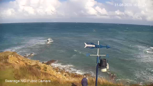 A view of a coastal scene from a webcam, showing the ocean with small sailboats in the distance under a partly cloudy sky. In the foreground, there is a person standing near a weather station or sensor mounted on a pole on a grassy, rocky cliff. The image has a timestamp in the upper right corner reading "31-08-2026 16:01:53" and a label in the lower left corner that says "Swanage NCI Public Camera."