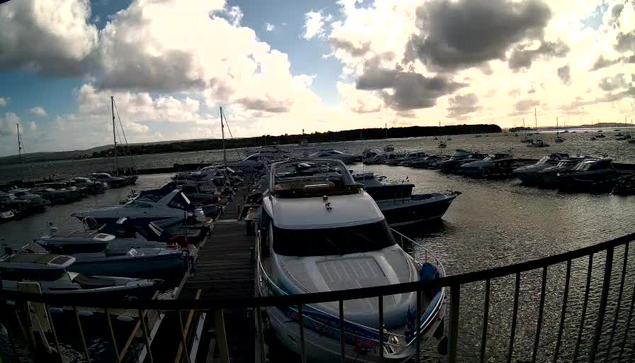 A marina filled with boats and yachts docked along wooden piers, with a partly cloudy sky and a distant shoreline in the background.