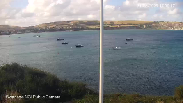 A view of a harbor with several boats floating on calm water. In the background, there are rolling hills with patches of greenery and cliffs along the shoreline. The sky is partly cloudy with some sunlight filtering through. The image appears to be split into two sections, possibly showing a comparison from a side-by-side camera setup. The timestamp in the top right corner indicates it was taken on August 8, 2023, at 5:00 PM. The bottom left corner has text that reads "Swanage NCI Public Camera."