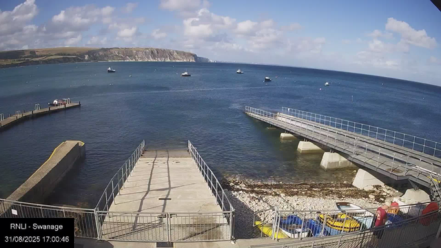 A view of a coastal scene with a small harbor, featuring two concrete piers extending into the water. The left pier is shorter, and the right pier is longer with a metal railing on top. Several small boats are anchored in the water. In the foreground, there is a rocky shoreline with colorful kayaks and paddle boats stored in a fenced area. The background shows a cliffside landscape under a partly cloudy sky.