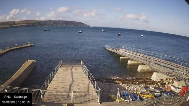 A view from a webcam showing a small harbor with a concrete pier extending into calm blue water. Several boats are floating in the distance, and a larger island or landmass with cliffs is visible on the horizon under partly cloudy skies. There is a fenced area in the foreground, with a few parked vehicles and some equipment. The timestamp reads "31/08/2025 18:00:16" and the location is noted as "RNLI - Swannage."