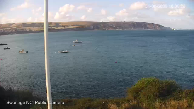 A view of a coastal scene with several small boats floating on calm blue water. In the background, there are white cliffs and a partly cloudy sky. A pole partially obstructs the view on the left side, and some green bushes are visible at the bottom right corner. The timestamp in the upper right corner indicates the date and time as 31-08-2026 18:00:19. The bottom left corner has text that reads "Swanage NCI Public Camera."
