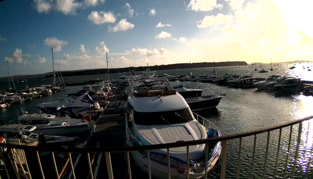 A marina filled with various boats and yachts docked along the pier, with the sun setting over the water and a partly cloudy sky in the background. The image is taken from a viewpoint behind a curved railing.