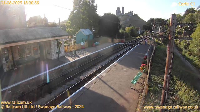 A small train station platform is shown in the image with railway tracks running alongside. The platform has a green bench, some potted flowers, and a small light green shed. There are tall wooden utility poles with electrical wires along the right side of the tracks. In the background, a hill with ruins or a castle is visible, surrounded by trees and greenery. The sky is clear, and the sun is shining, casting shadows on the platform. The timestamp indicates the image was taken on August 31, 2025, at 6:00 PM.