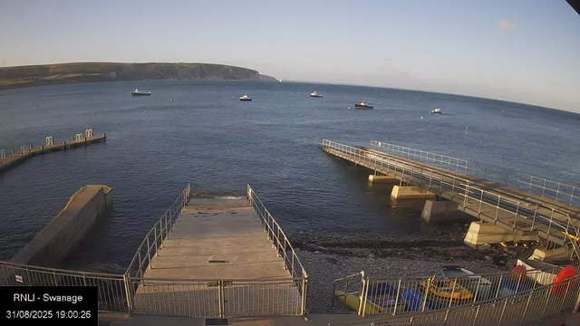 A seaside view showing a calm harbor with a concrete pier extending into the water, a walkway with metal railings on either side, and several small boats anchored in the distance. There is a rugged shoreline with rocks at the base of the pier. The background features a large headland or cliffs along the horizon, and the sky is mostly clear with a few small clouds.