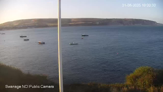 A split-screen image showing two views of a coastal scene. The left side displays a slightly different angle of the same scene, with a few small boats floating on the water near the shore, and a hillside covered in grass and sparse vegetation in the foreground. The right side shows a broader view of the water with several boats, including a larger vessel, and a distant coastline with cliffs. The timestamp in the top right corner indicates the date and time as August 31, 2025, at 13:00:30. The bottom left corner identifies the source as "Swanage NCI Public Camera."