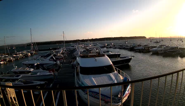 A marina filled with numerous boats and yachts docked along wooden piers, with a calm water surface reflecting the sky. The sky shows the warm glow of a setting or rising sun near the horizon.