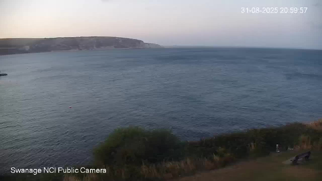 A wide view of a calm ocean with gentle waves, a distant coastline with cliffs, and a faint evening sky with soft colors. There are some trees and a bench visible in the lower right corner. The timestamp in the top right corner shows the date and time as 31-08-2025 20:59:57.